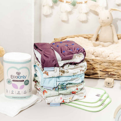 Stack of baby diapers with an EcoKindly container on a white surface, surrounded by baby toys and items.